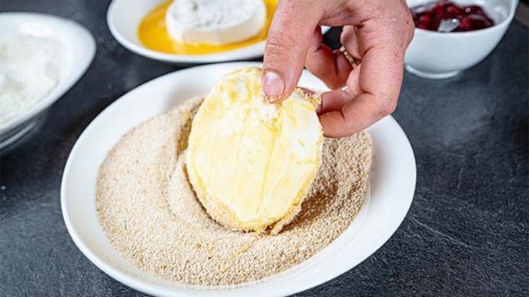 Eine Hand wendet ein mit Ei bedecktes Stück Camembert in Brotkrümeln, weitere Zutaten wie Ei und Beilage sind im Hintergrund.