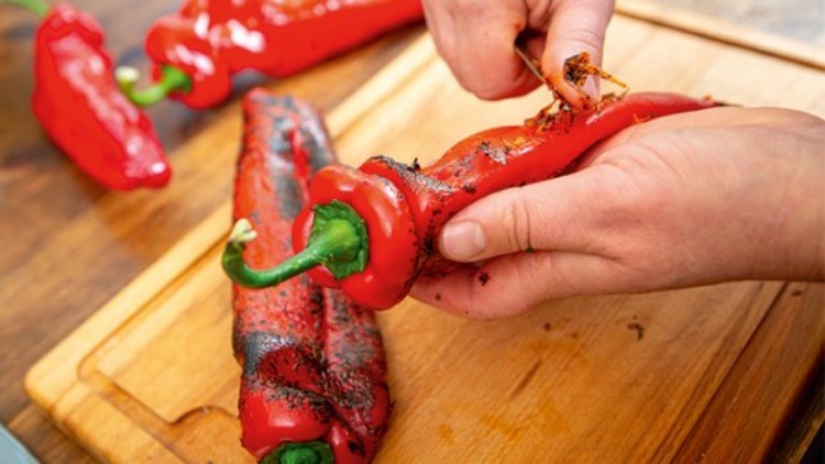 Geröstete rote Paprika werden auf einem Holzbrett geschält, im Hintergrund liegen weitere Paprika.