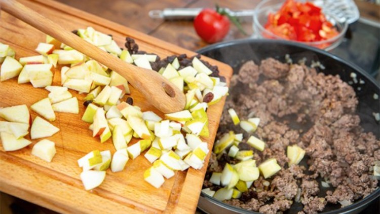 Holzbrett mit geschnittenen Äpfeln und Rosinen neben einer Pfanne mit gebratenem Hackfleisch, im Hintergrund Paprikawürfel.