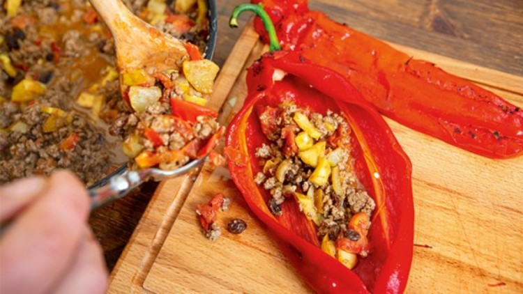 Eine rote Paprika wird mit gebratenem Hackfleisch und Gemüse aus einer Pfanne gefüllt, daneben liegt eine halbierte Paprika.
