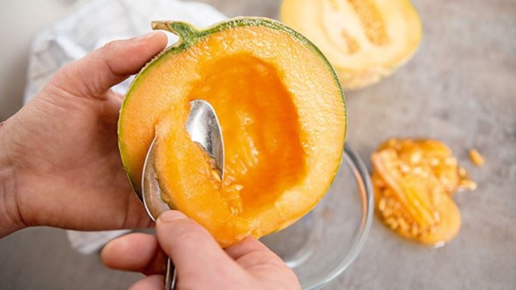 Eine halbierte Melone wird mit einem Löffel ausgehöhlt, die Kerne liegen daneben auf einer Schale.