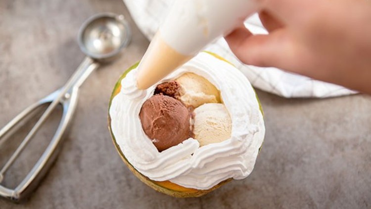 Eine halbierte Melone mit Kugeln Eis und Sahne, daneben ein Eiskugelportionierer auf grauem Untergrund.