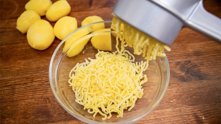 Gekochte Kartoffeln werden mit einer silbernen Kartoffelpresse in eine Glasschüssel auf einem Holztisch gepresst.