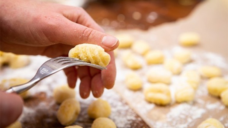 Eine Hand formt mit einer Gabel Gnocchi aus Teig, im Hintergrund liegen weitere Teigstücke auf bemehltem Backpapier.