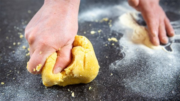 Eine Hand knetet gelben Teig auf einer bemehlten dunklen Arbeitsfläche, daneben liegt etwas weiteres Mehl.