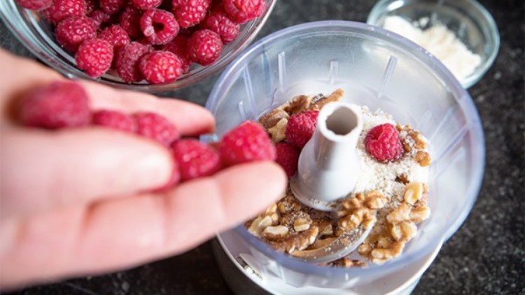 Eine Hand hält Himbeeren über einen Mixer, der mit Walnüssen, Himbeeren und gemahlenen Mandeln befüllt ist.