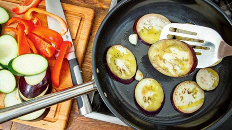 In einer schwarzen Pfanne werden Auberginenscheiben angebraten, daneben liegen geschnittene Zucchini und Paprika auf einem Holzbrett.
