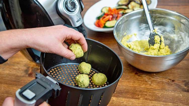 Eine Person legt grüne Teigbällchen in die Schale einer Heißluftfritteuse, daneben eine Schüssel mit Teig und ein Teller mit Gemüse.