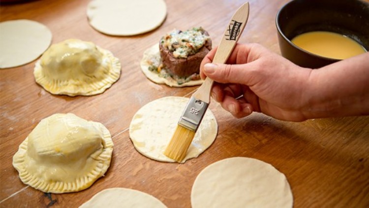 Eine Hand bestreicht runde Teiglinge mit einem Pinsel, daneben gefüllte Teigtaschen und eine Schüssel mit gelber Flüssigkeit.