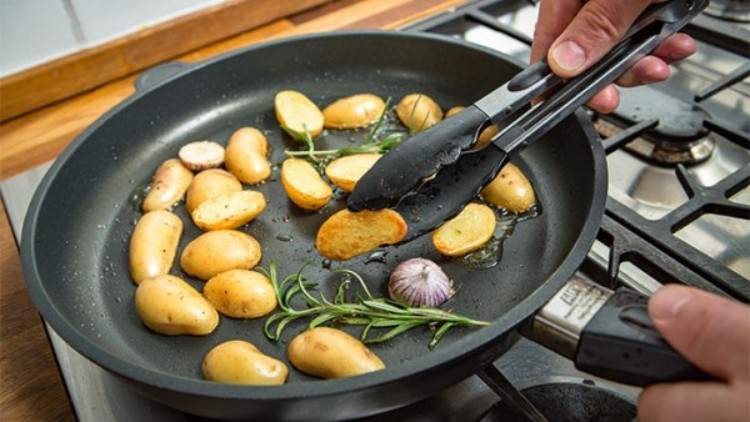Ganze und halbierte Kartoffeln werden mit einer Zange in einer Pfanne mit Knoblauch, Öl und Rosmarin auf einem Herd gewendet.