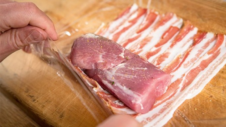 Ein Stück rohes Schweinefleisch wird in eine Schicht aus dünnem, rohem Speck auf einem Holzbrett eingewickelt.
