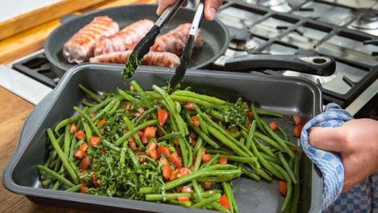 Grüne Bohnen mit gehackten Tomaten und Kräutern in einer schwarzen Auflaufform, im Hintergrund Pfanne mit Fleischrollen.