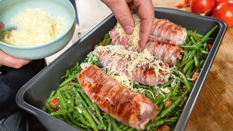 Eine Hand streut geriebenen Käse auf vier mit Speck umwickelte Fleischstücke in einer Auflaufform mit grünen Bohnen.