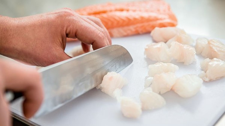 Eine Hand schneidet mit einem Messer weiße Fischstücke auf einem Schneidebrett, im Hintergrund liegt ein Lachsfilet.