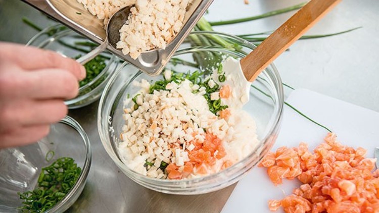 Eine Glasschüssel mit Joghurt, Kräutern, gewürfeltem Lachs und Blumenkohl wird mit weiteren Blumenkohlstücken befüllt.