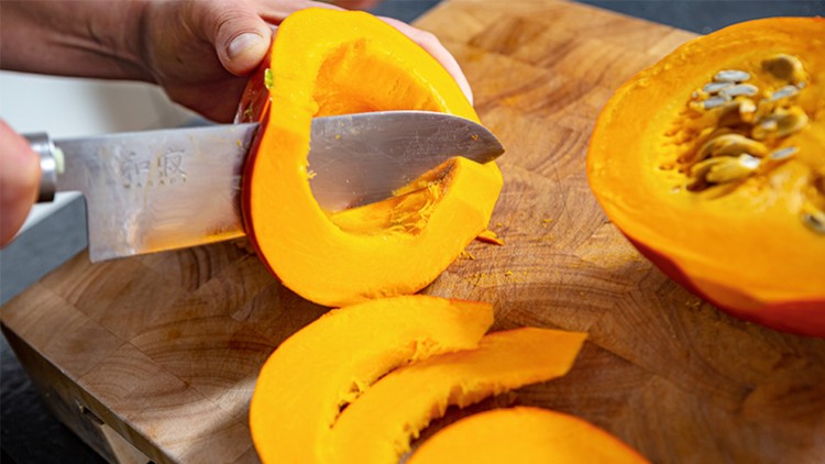 Ein Messer schneidet einen orangefarbenen Kürbis auf einem hölzernen Schneidebrett in Stücke.