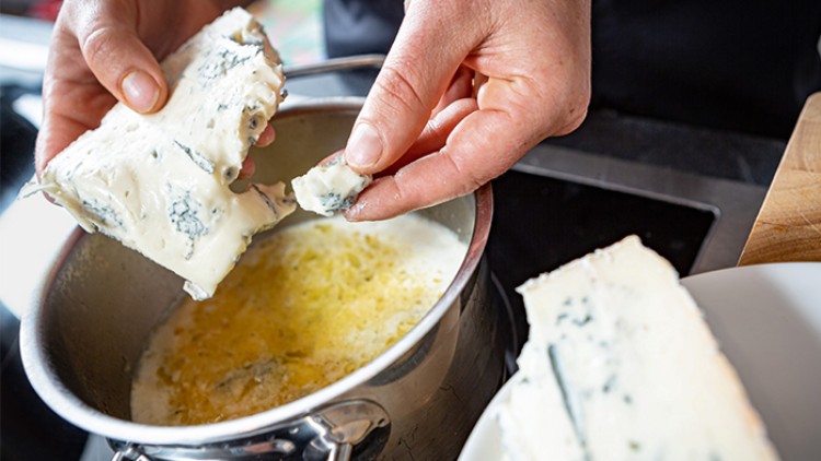Ein Stück Blauschimmelkäse wird mit der Hand gebrochen und in einen Topf mit geschmolzener Butter gegeben.