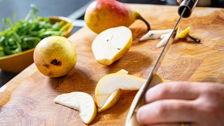 Eine Hand schneidet eine gelbe Birne auf einem Holzbrett, daneben liegen zwei Birnen und eine Schale mit grünem Salat.