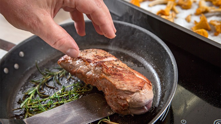 Ein gebratenes Steak liegt in einer Pfanne mit frischem Rosmarin, daneben ist eine Hand und ein Backblech mit Pfifferlingen.