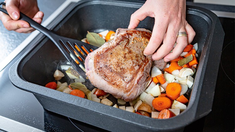 Ein Braten wird mit einem schwarzen Pfannenwender auf einem Bett aus Gemüse wie Karotten, Zwiebeln und Lorbeerblättern gewendet.