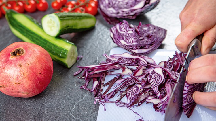 Ein aufgeschnittener Granatapfel, Gurken, Tomaten und Rotkohl, davon wird Rotkohl auf einem Brett mit einem Messer geschnitten.