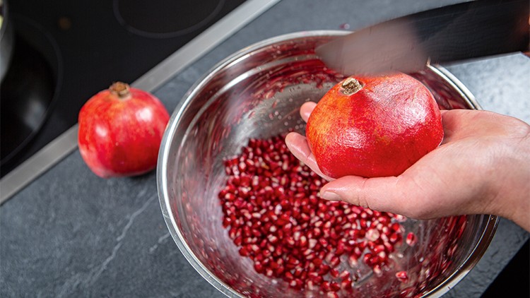 Eine Hand hält einen Granatapfel über eine Schüssel mit Granatapfelkernen, während ein Messer den Granatapfel schneidet.