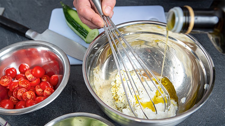 Eine Hand hält einen Schneebesen in einer Metallschüssel mit Joghurt, Gewürzen und Öl, daneben Tomaten und Gurken auf einem Brett.