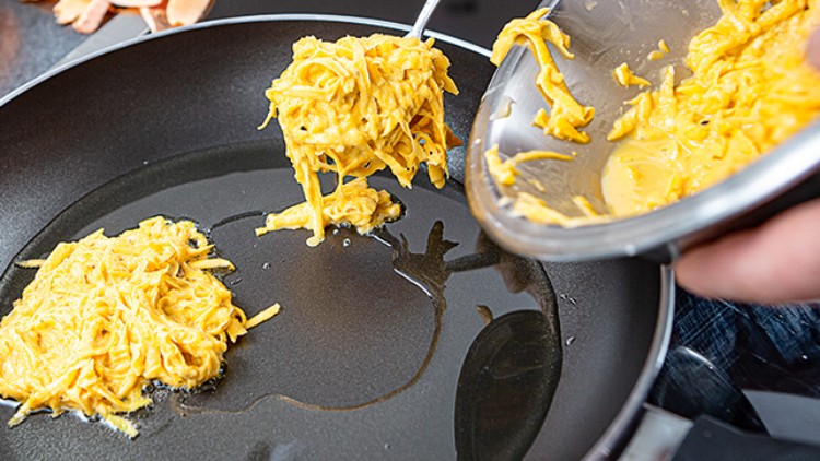 Zwei Portionen gelber Teigmasse werden mit einem Löffel aus einer Schüssel in eine schwarze Pfanne mit Öl gegeben.