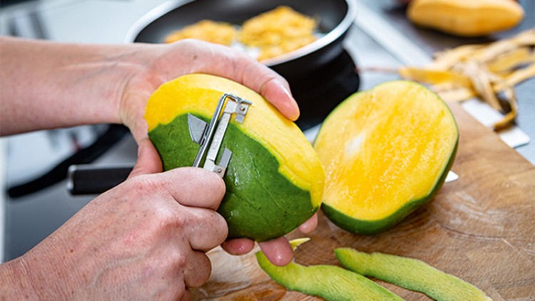 Zwei Hände schälen mit einem Sparschäler eine reife grüngelbe Mango, im Hintergrund liegen Schalen und eine Pfanne.