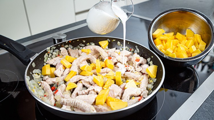 In einer Pfanne mit angebratenem Putenfleisch, Zwiebeln und Paprika wird Sahne aus einem Glaskännchen hinzugefügt.