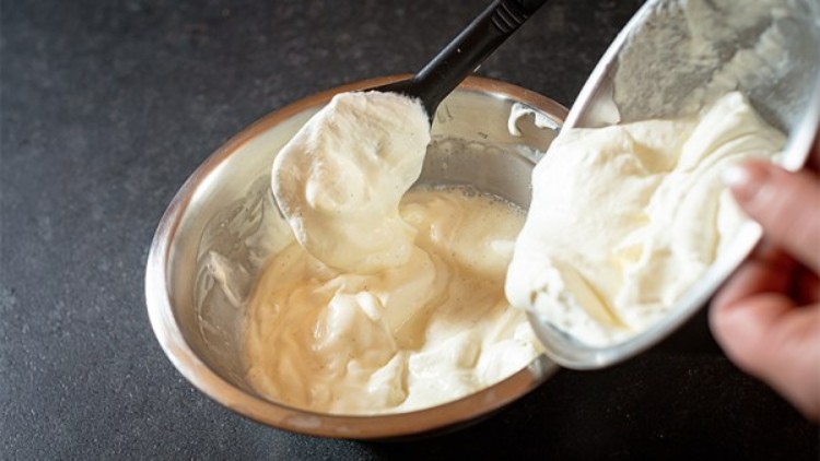 Metallschüssel mit heller Creme, links ein schwarzer Löffel, rechts wird Creme aus einer Schale hinzugefügt.