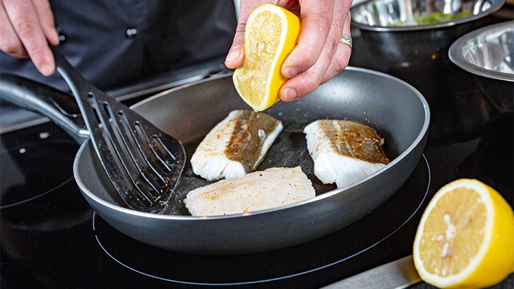 Drei Stücke Fisch werden in einer Pfanne gebraten, während eine halbe Zitrone über dem Fisch ausgedrückt wird.