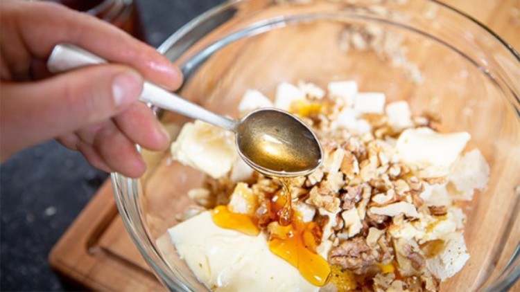 Eine Hand hält einen Löffel mit Honig über einer Glasschüssel mit Walnüssen, Butter und weißen Würfeln.