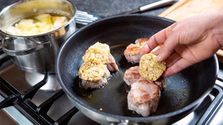 Hand legt eine Semmelknödelscheibe auf angebratene Schweinefiletstücke in einer Pfanne, daneben ein Topf mit Kartoffeln.