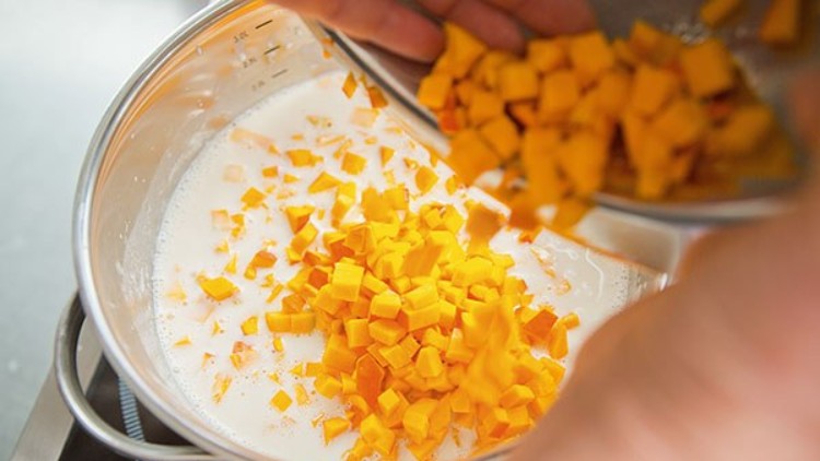 Kleine Würfel aus orangefarbenem Gemüse werden in einen Kochtopf mit heller Flüssigkeit gegeben.