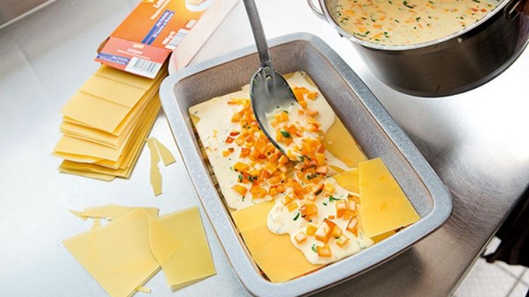Eine Auflaufform wird mit Lasagneblättern, Soße und Gemüse befüllt, daneben liegen weitere Teigplatten und eine Verpackung.