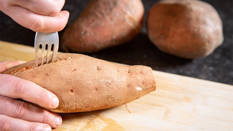 Eine Hand sticht mit einer Gabel Löcher in eine rohe Süßkartoffel auf einem Holzbrett, weitere Süßkartoffeln liegen im Hintergrund.