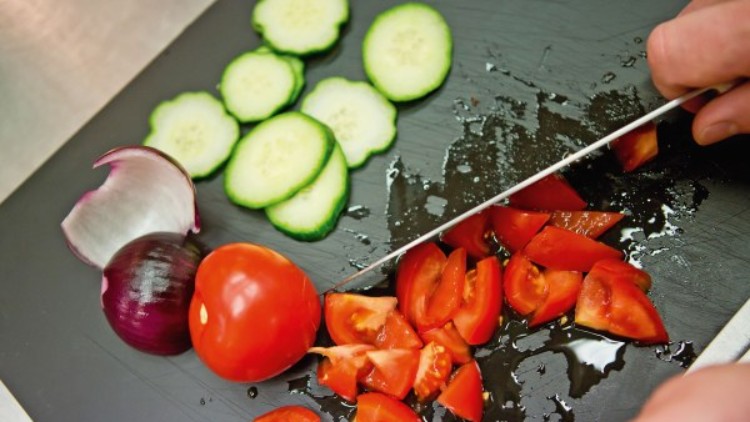 Auf einem Schneidebrett werden eine Tomate geschnitten, daneben liegen Gurkenscheiben und eine angeschnittene rote Zwiebel.