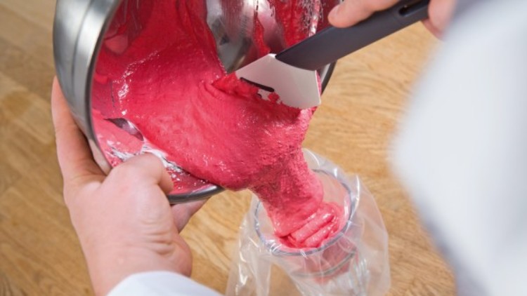 Rosa Teigmasse wird aus einer Metallschüssel mit einem Spatel in einen Einwegspritzbeutel über einem Glas gefüllt.