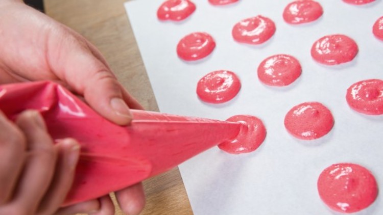 Eine Hand drückt rosafarbenen Teig aus einem Spritzbeutel auf ein Backpapier auf einem Holztisch.