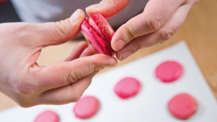 Zwei Hände drücken eine rote, gefüllte Macaronhälfte zusammen, im Hintergrund liegen weitere Macarons auf Backpapier.
