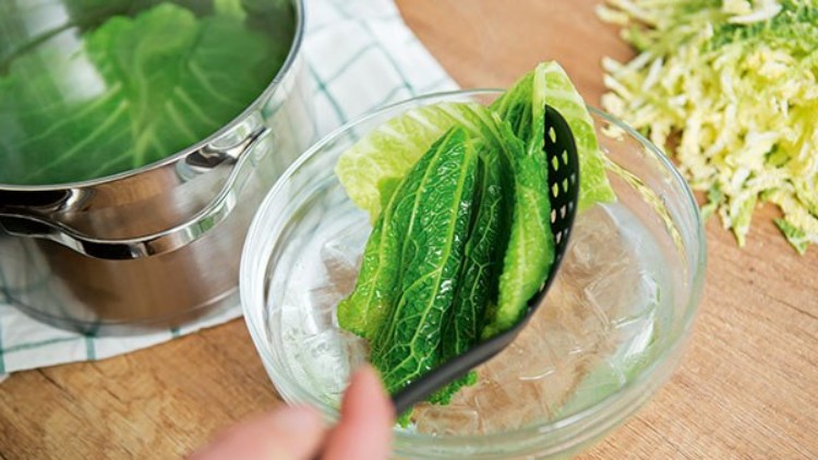 Grüne Kohlblätter werden mit einem schwarzen Schaumlöffel aus einer Glasschüssel mit Eiswasser gehoben, daneben ein Topf und geriebener Kohl.