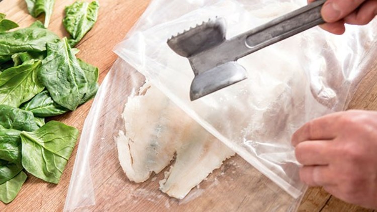 Ein Fleischklopfer liegt auf einem weißen Fischfilet in einem Plastikbeutel, daneben frische grüne Spinatblätter auf Holzuntergrund.