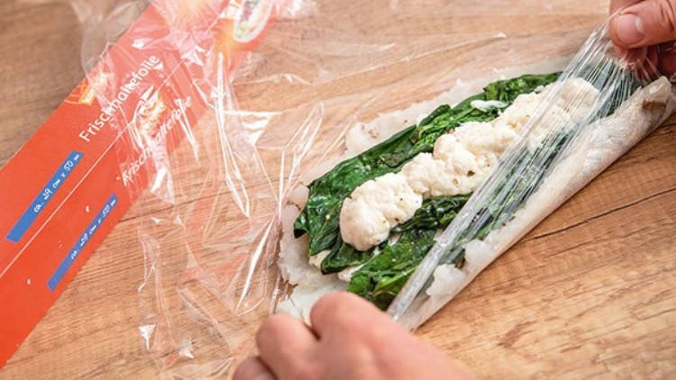 Hände wickeln Fischfilet mit Spinat und weißer Füllung in Frischhaltefolie, daneben liegt eine rote Verpackung mit Aufschrift.