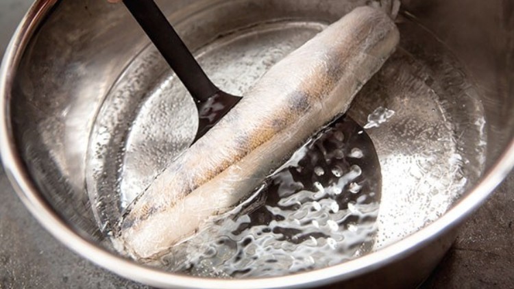 Eine eingeschweißte Wurst wird mit einem schwarzen Pfannenwender in einem Topf mit kochendem Wasser gehalten.