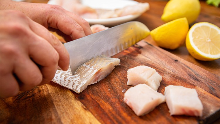 Hände schneiden mit einem Messer weißen Fisch auf einem Holzbrett, daneben liegen ganze und halbe gelbe Zitronen.