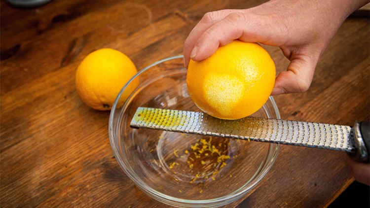 Hand reibt mit einer Metallreibe die Schale einer Orange ab, darüber eine Glasschüssel auf einem Holztisch.