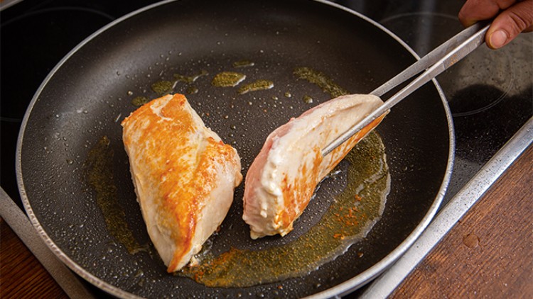 Zwei Hähnchenbrustfilets werden in einer Pfanne mit etwas Öl angebraten, eines wird mit einer Zange gewendet.