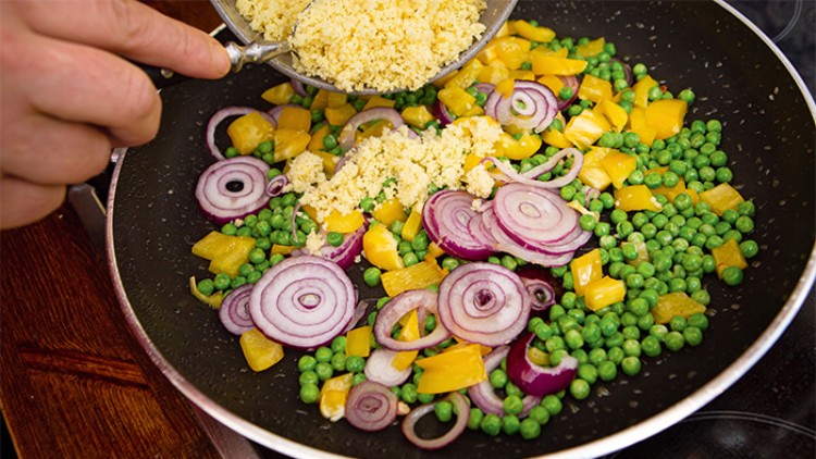 In einer Pfanne liegen grüne Erbsen, gelbe Paprikastücke und rote Zwiebelringe, darüber wird Couscous hinzugegeben.