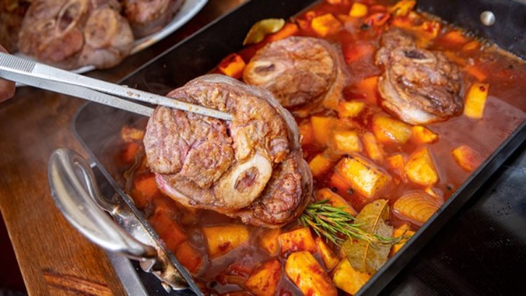 Gekochtes Fleischstück wird mit einer Zange aus einer schwarzen Pfanne mit rotem Eintopf, Kartoffeln und Kräutern gehoben.